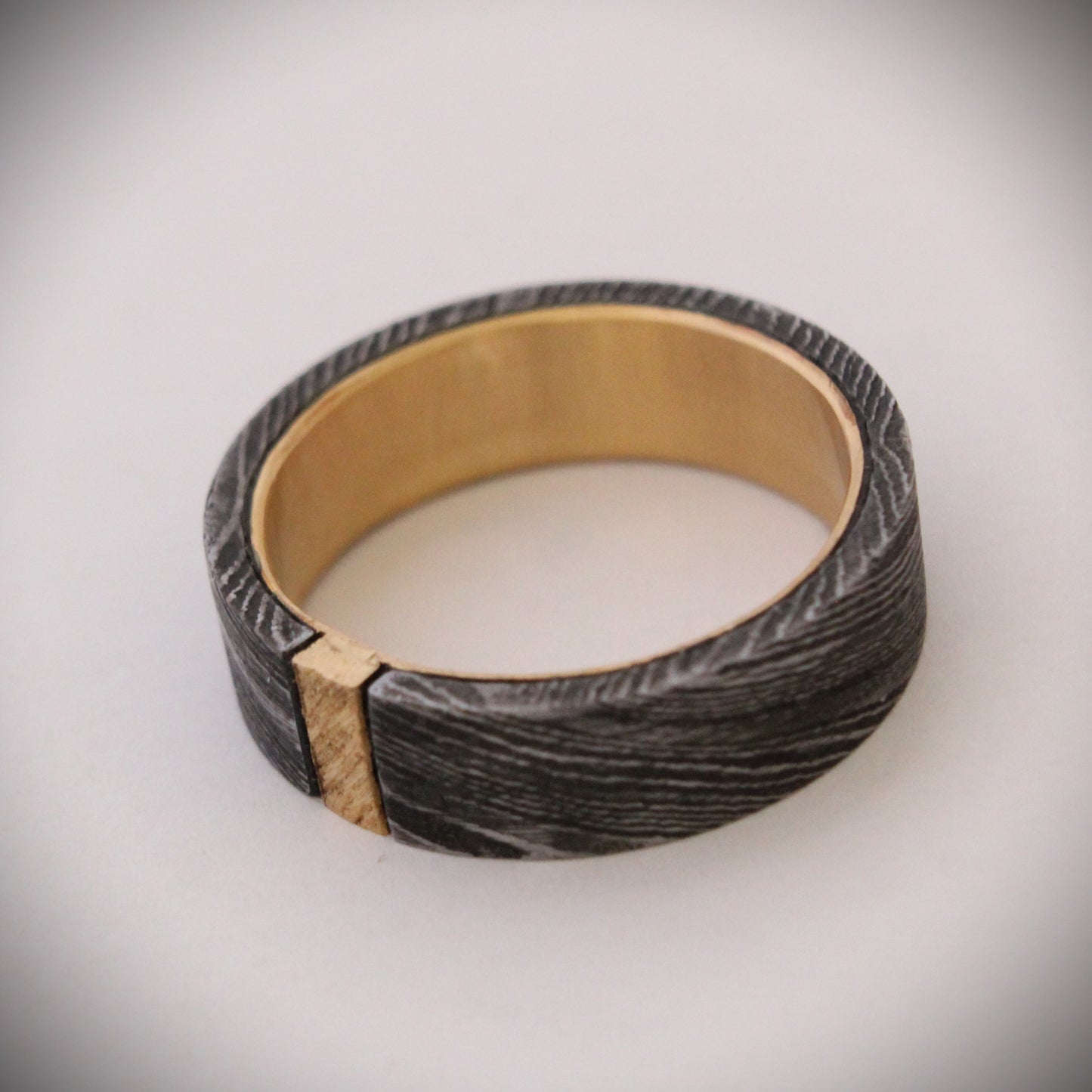 Damascus steel ring with 18ct yellow gold inner band and patterned gold accent: A striking ring featuring a rugged outer band of authentic Damascus steel with intricate swirling patterns, paired with a luxurious 18ct yellow gold inner band. A delicate patterned gold accent runs along the outer edge, enhancing the contrast between the steel’s dark, fluid designs and the warm glow of the gold, creating a unique, elegant look. Ideal as a wedding band, engagement ring, or statement piece.
