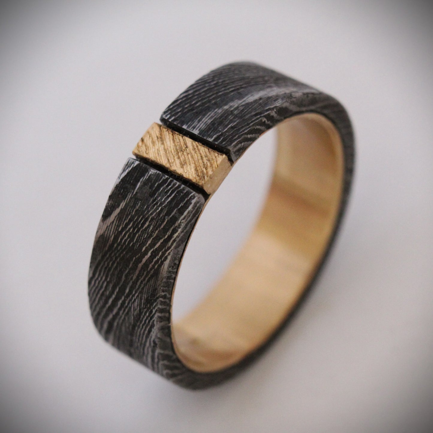 Damascus steel ring with 18ct yellow gold inner band and patterned gold accent: A striking ring featuring a rugged outer band of authentic Damascus steel with intricate swirling patterns, paired with a luxurious 18ct yellow gold inner band. A delicate patterned gold accent runs along the outer edge, enhancing the contrast between the steel’s dark, fluid designs and the warm glow of the gold, creating a unique, elegant look. Ideal as a wedding band, engagement ring, or statement piece.