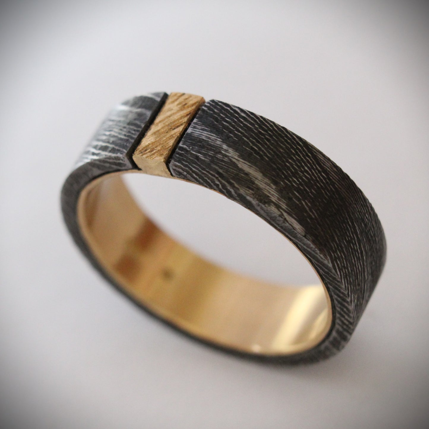 Damascus steel ring with 18ct yellow gold inner band and patterned gold accent: A striking ring featuring a rugged outer band of authentic Damascus steel with intricate swirling patterns, paired with a luxurious 18ct yellow gold inner band. A delicate patterned gold accent runs along the outer edge, enhancing the contrast between the steel’s dark, fluid designs and the warm glow of the gold, creating a unique, elegant look. Ideal as a wedding band, engagement ring, or statement piece.