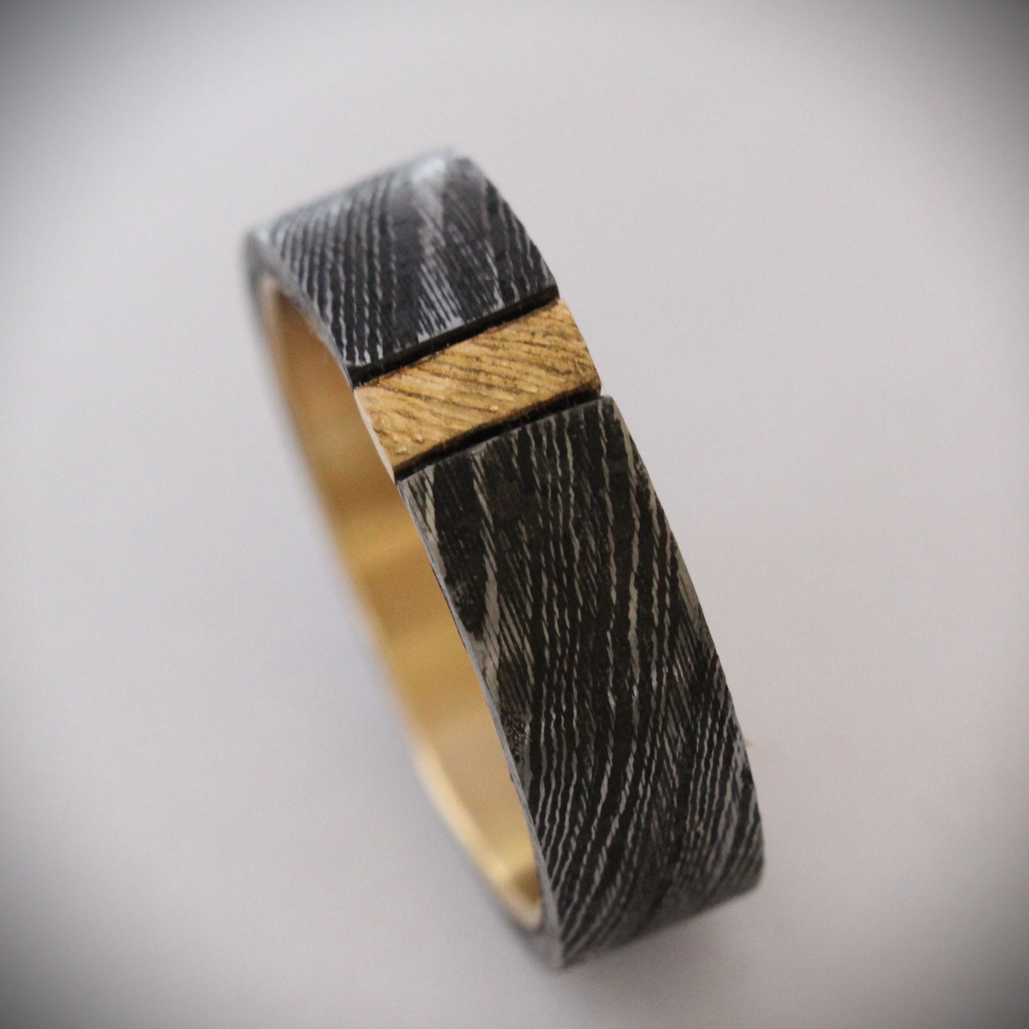 Damascus steel ring with 18ct yellow gold inner band and patterned gold accent: A striking ring featuring a rugged outer band of authentic Damascus steel with intricate swirling patterns, paired with a luxurious 18ct yellow gold inner band. A delicate patterned gold accent runs along the outer edge, enhancing the contrast between the steel’s dark, fluid designs and the warm glow of the gold, creating a unique, elegant look. Ideal as a wedding band, engagement ring, or statement piece.