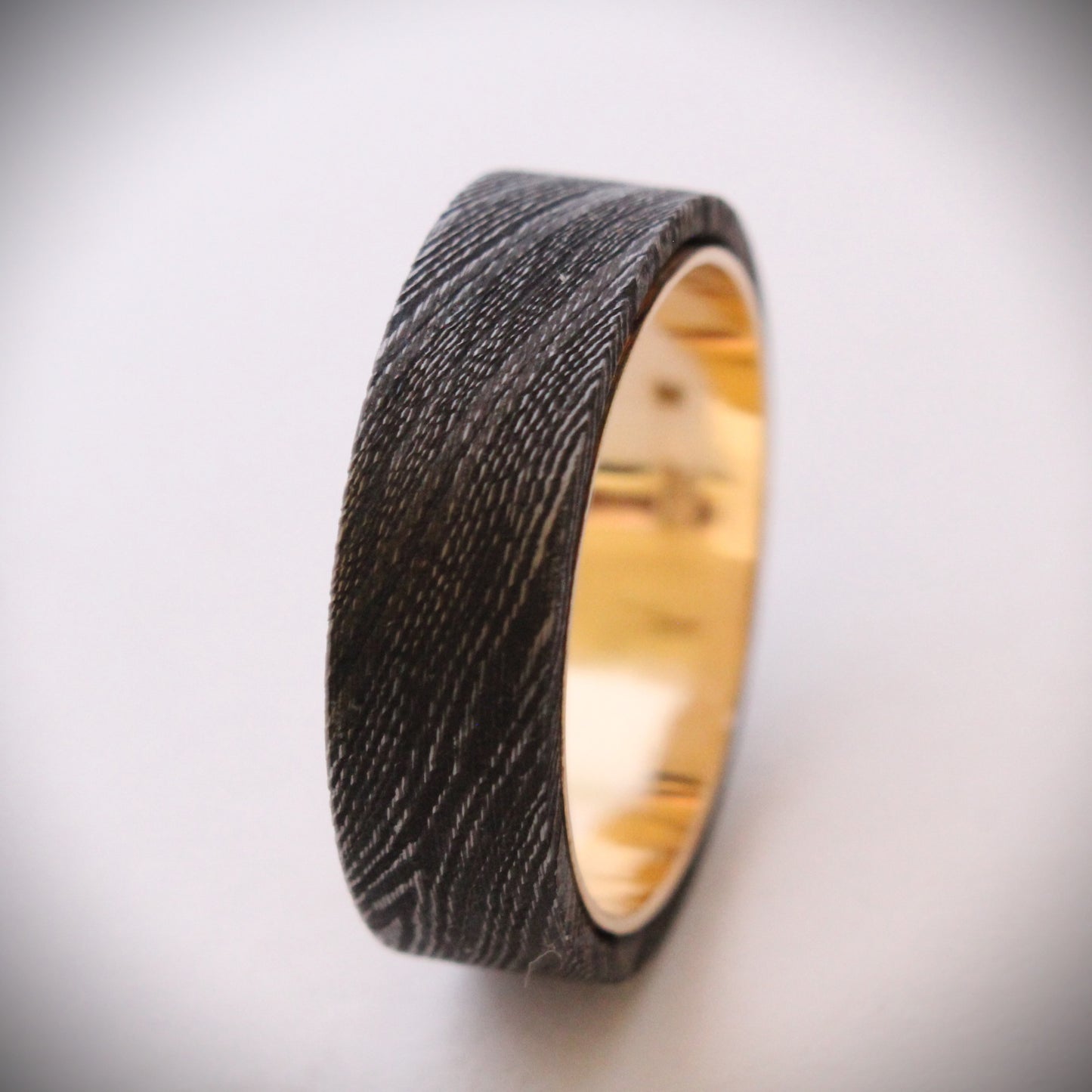 A unique ring featuring a textured exterior of Damascus steel, showcasing intricate swirling patterns, complemented by a smooth inner band made of 18ct yellow gold. The contrast between the dark steel and warm gold creates a striking visual effect, making it a standout piece of jewelry suitable for both everyday wear and special occasions.