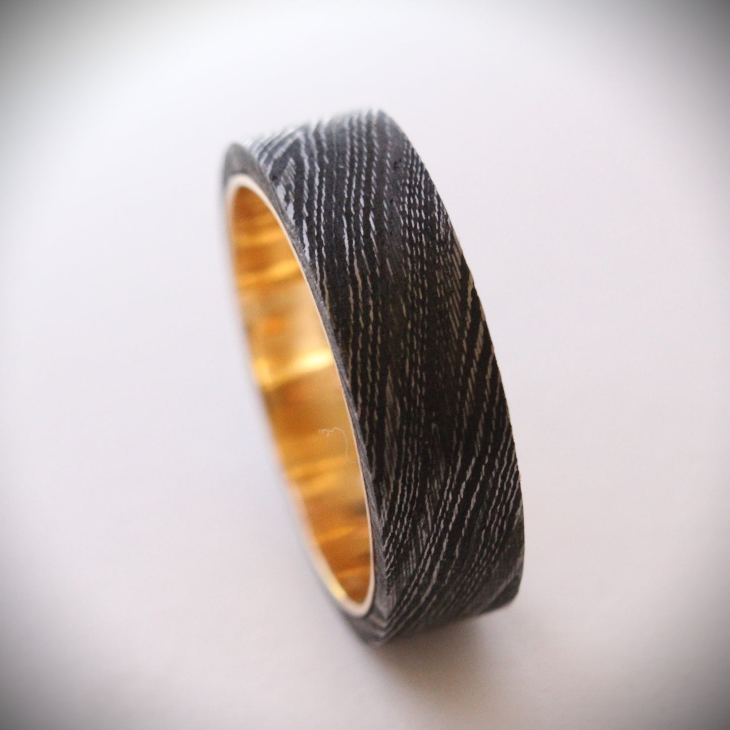A unique ring featuring a textured exterior of Damascus steel, showcasing intricate swirling patterns, complemented by a smooth inner band made of 18ct yellow gold. The contrast between the dark steel and warm gold creates a striking visual effect, making it a standout piece of jewelry suitable for both everyday wear and special occasions.