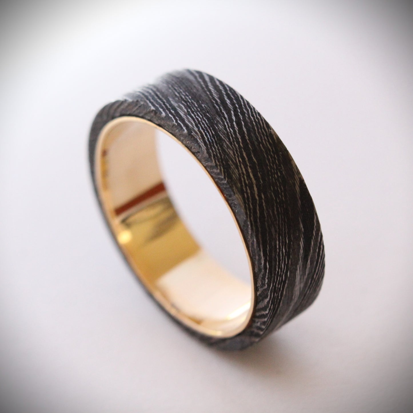 A unique ring featuring a textured exterior of Damascus steel, showcasing intricate swirling patterns, complemented by a smooth inner band made of 18ct yellow gold. The contrast between the dark steel and warm gold creates a striking visual effect, making it a standout piece of jewelry suitable for both everyday wear and special occasions.