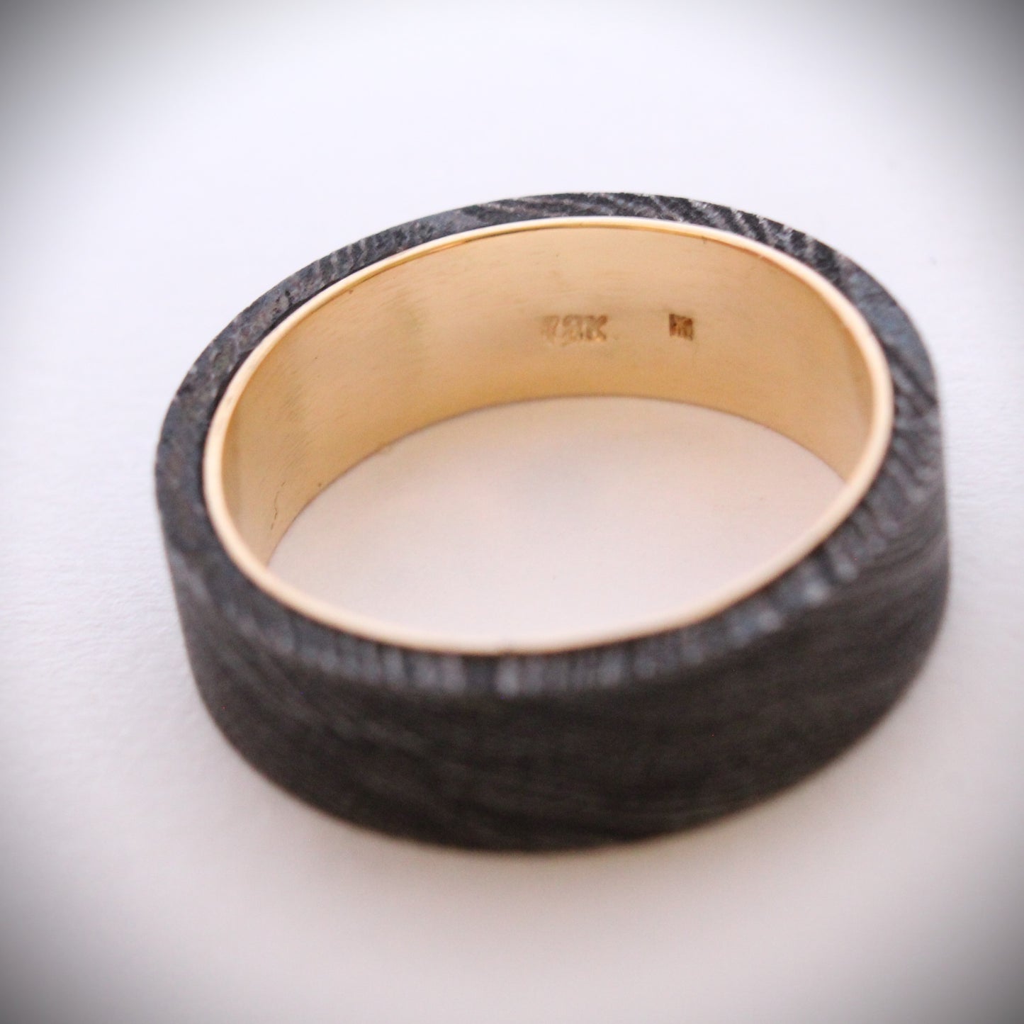A unique ring featuring a textured exterior of Damascus steel, showcasing intricate swirling patterns, complemented by a smooth inner band made of 18ct yellow gold. The contrast between the dark steel and warm gold creates a striking visual effect, making it a standout piece of jewelry suitable for both everyday wear and special occasions.