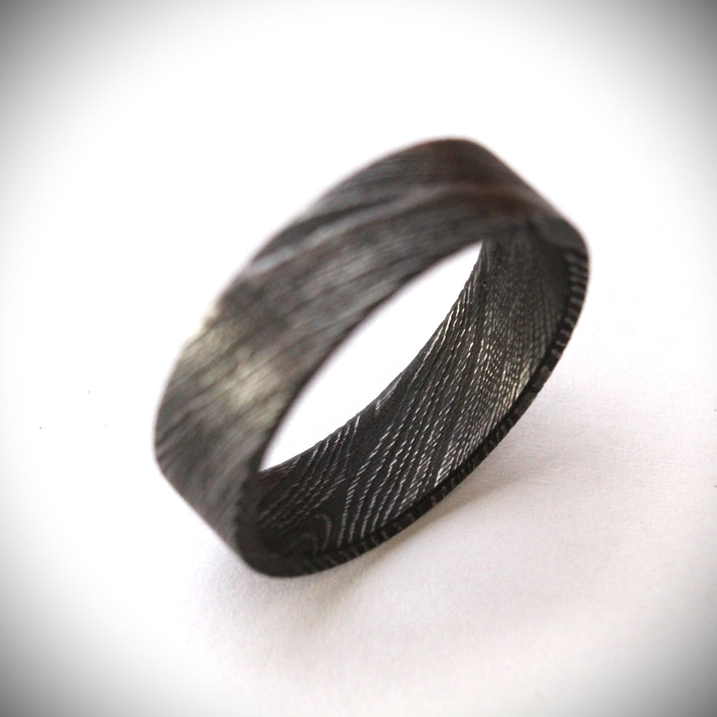 Stunning Damascus steel ring with intricate flowing patterns, heat-treated and blackened for a bold, modern look. The outer band features a unique, layered design with a dark, matte finish, combining strength and elegance in a one-of-a-kind piece of fine craftsmanship. Perfect for a wedding band, engagement ring, or statement piece.