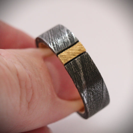 Damascus steel ring with 18ct yellow gold inner band and patterned gold accent: A striking ring featuring a rugged outer band of authentic Damascus steel with intricate swirling patterns, paired with a luxurious 18ct yellow gold inner band. A delicate patterned gold accent runs along the outer edge, enhancing the contrast between the steel’s dark, fluid designs and the warm glow of the gold, creating a unique, elegant look. Ideal as a wedding band, engagement ring, or statement piece.