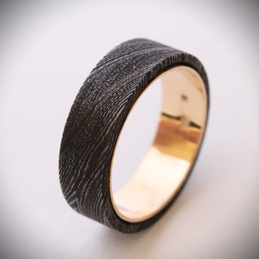 A unique ring featuring a textured exterior of Damascus steel, showcasing intricate swirling patterns, complemented by a smooth inner band made of 18ct yellow gold. The contrast between the dark steel and warm gold creates a striking visual effect, making it a standout piece of jewelry suitable for both everyday wear and special occasions.
